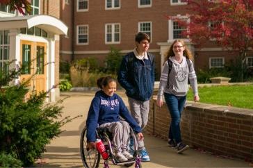 三个主要研究学生, one in a wheelchair and two standing alongside her, walk and wheel in the courtyard at 主要研究's Murkland Hall.